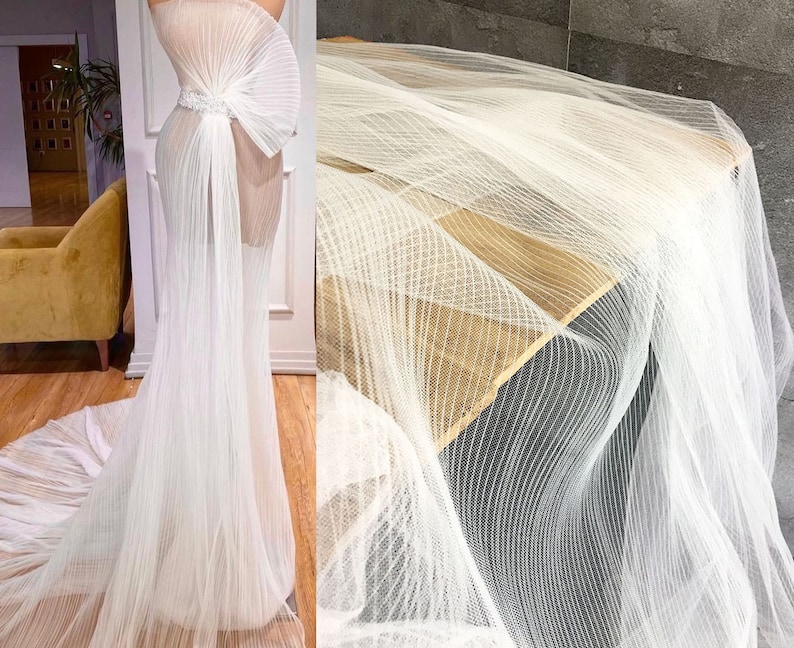 May include: A white wedding dress with a sheer, pleated bodice and a flowing skirt. The dress is made of a delicate, lightweight fabric that drapes beautifully. The skirt is pleated and has a long train.