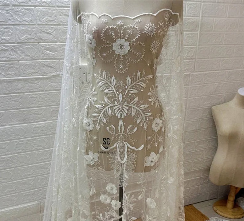 May include: White beaded lace fabric with floral designs and a scalloped edge. The fabric is displayed on a white mannequin.