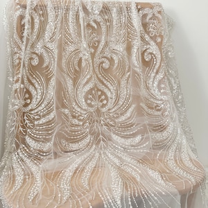 Puede incluir: Tela de tul blanca con intrincados patrones florales y de volutas con cuentas y lentejuelas. El delicado diseño está drapeado sobre una silla de madera, mostrando la textura de la tela. Ideal para ropa de novia o formal.