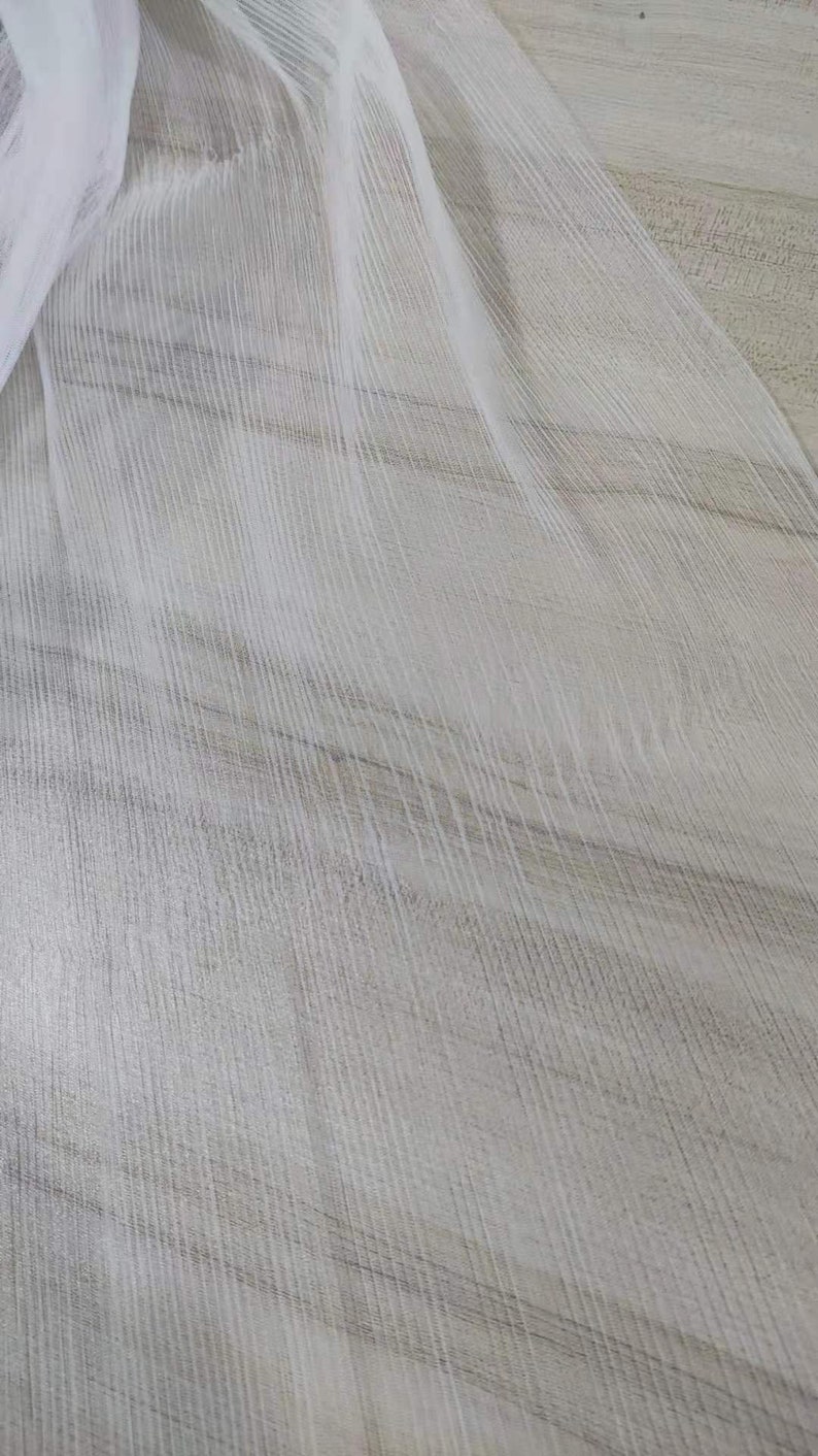May include: White sheer fabric with a textured, striped pattern. The fabric is draped over a light brown surface.