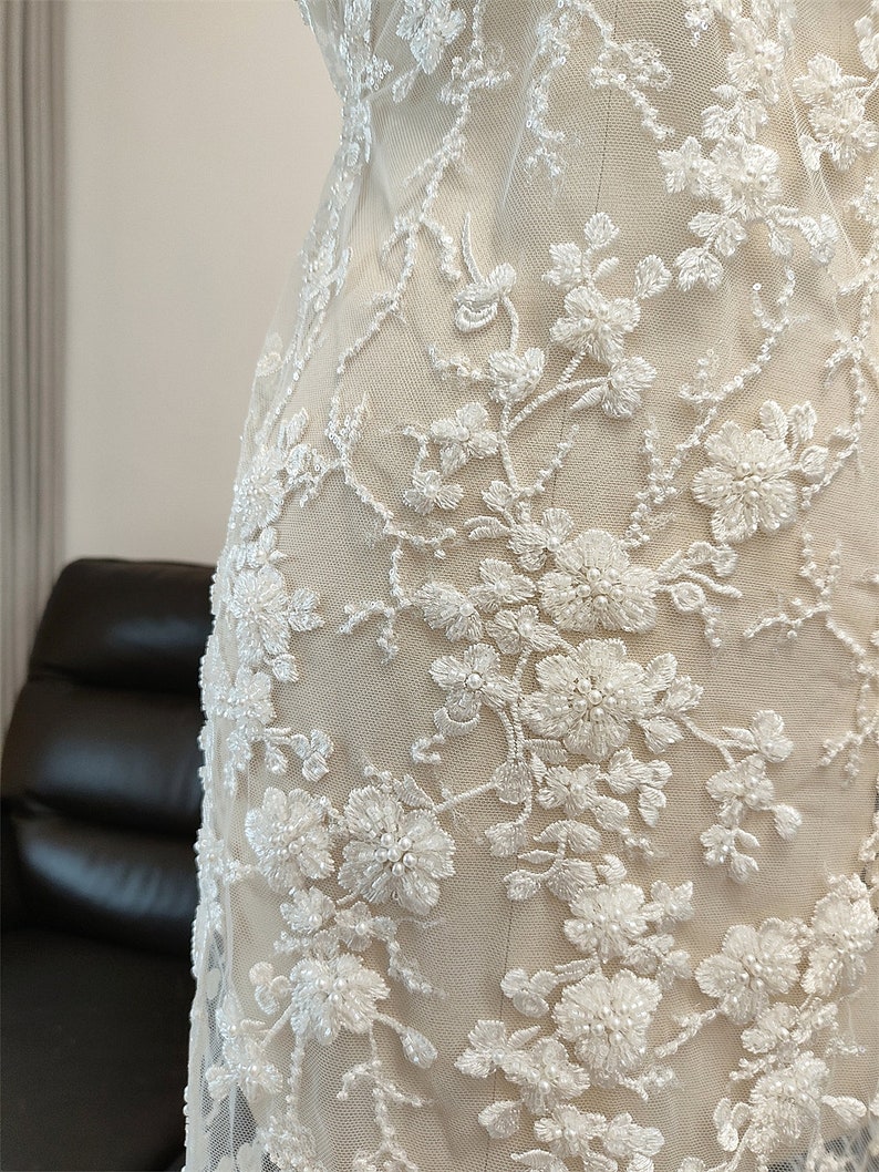May include: Close-up of ivory fabric with intricate white floral embroidery. The design features detailed, three-dimensional flowers and beaded accents, creating a textured and elegant appearance. The fabric is likely used for bridal or formal wear.