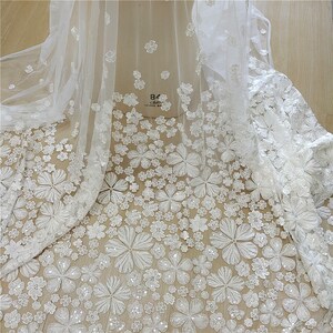 May include: White floral lace fabric with a delicate pattern of small and large flowers. The fabric has a sheer, lightweight texture and is perfect for wedding dresses, bridal veils, and other special occasion garments.