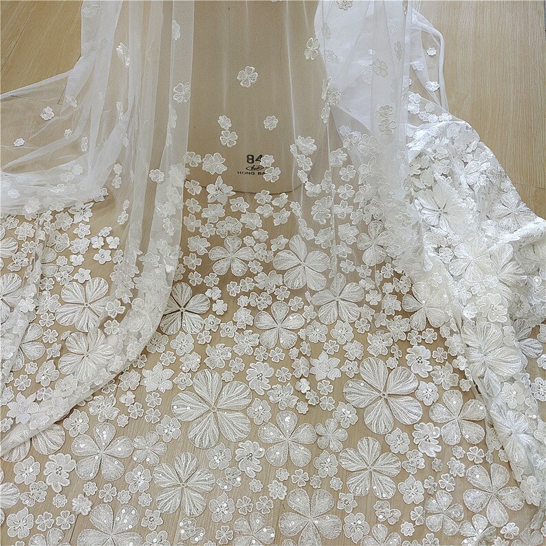 May include: White floral lace fabric with a delicate pattern of small and large flowers. The fabric has a sheer, lightweight texture and is perfect for wedding dresses, bridal veils, and other special occasion garments.