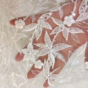May include: White floral lace fabric with a delicate pattern of leaves and flowers. The fabric is embellished with small, white sequins.