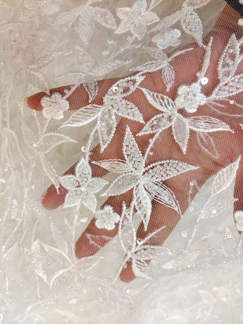 May include: White floral lace fabric with a delicate pattern of leaves and flowers. The fabric is embellished with small, white sequins.