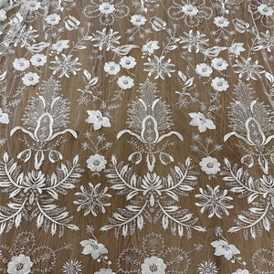 May include: White floral lace fabric with a delicate pattern of flowers and leaves. The lace is beaded with small, shiny beads and has a scalloped edge.