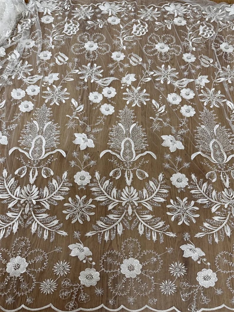 May include: White floral lace fabric with a delicate pattern of flowers and leaves. The lace is beaded with small, shiny beads and has a scalloped edge.