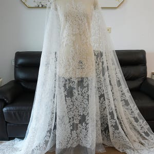 May include: White lace fabric draped over a mannequin. The lace features intricate floral patterns and is semi-sheer. The fabric is displayed against a neutral background, highlighting the delicate details of the lace.