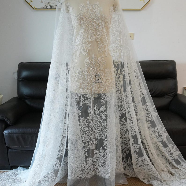 May include: White lace fabric draped over a mannequin. The lace features intricate floral patterns and is semi-sheer. The fabric is displayed against a neutral background, highlighting the delicate details of the lace.