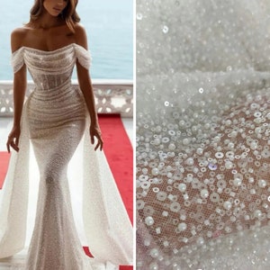 May include: A white off-the-shoulder wedding dress with a fitted bodice and a flowing skirt. The dress is embellished with sequins and beads, creating a shimmering effect. The fabric is sheer and delicate, and the dress has a train.