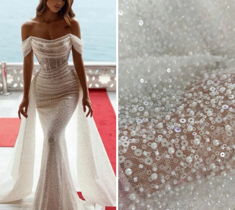 May include: A white off-the-shoulder wedding dress with a fitted bodice and a flowing skirt. The dress is embellished with sequins and beads, creating a shimmering effect. The fabric is sheer and delicate, and the dress has a train.