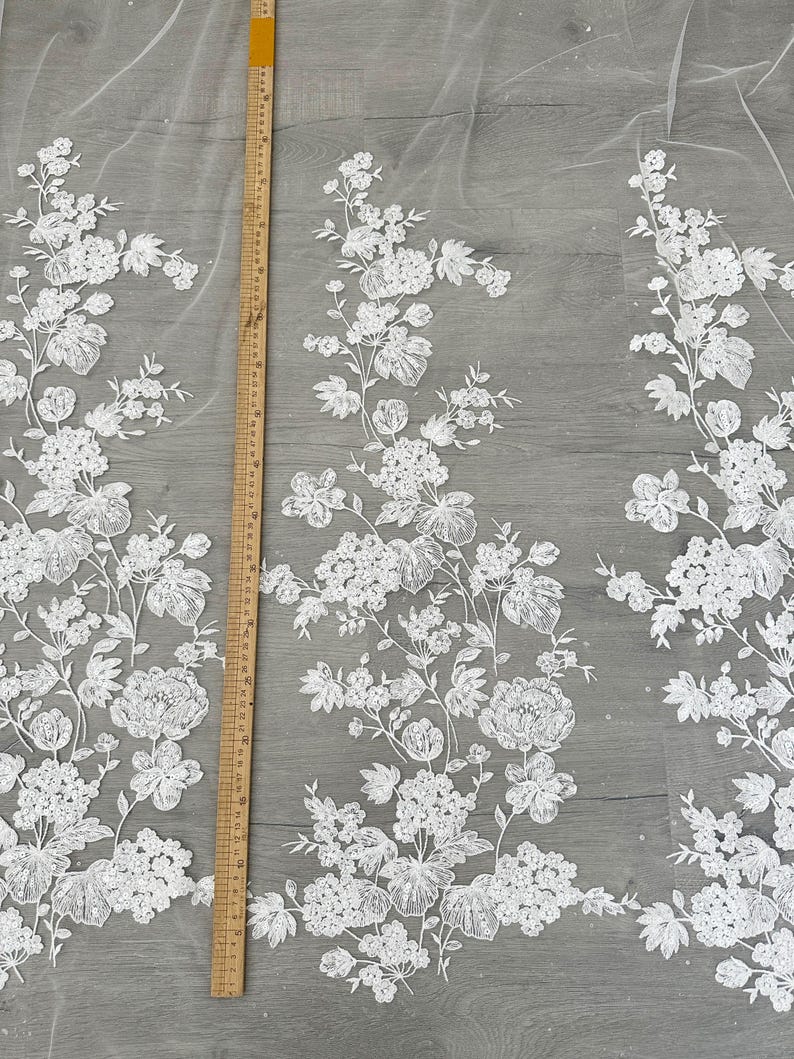 May include: White floral lace fabric with a ruler for scale. The lace features a delicate floral pattern with small white flowers and leaves. The fabric is sheer and lightweight, making it ideal for wedding dresses, bridal accessories, and other special occasion garments.
