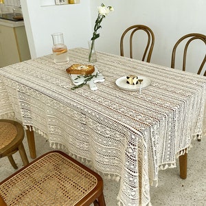 May include: A white crocheted tablecloth with fringe detailing covers a wooden table. The tablecloth has a delicate floral pattern and is set with a plate, a glass of water, and a piece of toast on a wooden cutting board.