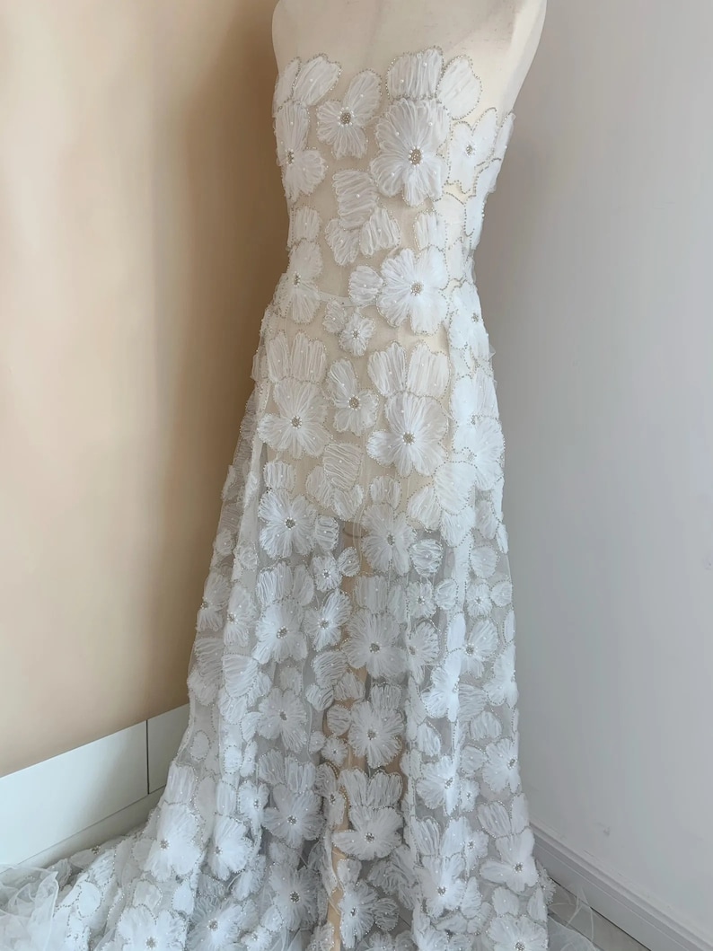 May include: White floral lace fabric with 3D flowers and a sheer background. The fabric is delicate and intricate, perfect for wedding dresses, evening gowns, or other special occasion garments.