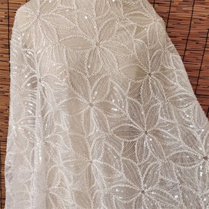 Peut inclure: Tissu blanc perl&eacute; avec un motif floral. Le tissu est fait de paillettes et de perles, et il a un aspect d&eacute;licat et f&eacute;minin.