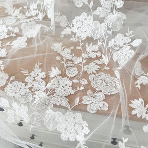 May include: White floral embroidered tulle fabric with a delicate pattern of flowers and leaves. The fabric is sheer and lightweight, making it ideal for wedding dresses, bridal veils, and other special occasion garments.