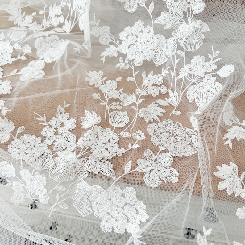 May include: White floral embroidered tulle fabric with a delicate pattern of flowers and leaves. The fabric is sheer and lightweight, making it ideal for wedding dresses, bridal veils, and other special occasion garments.