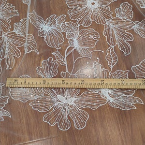 May include: White floral lace fabric with a beaded design. The fabric is laid out on a wooden surface with a ruler measuring 35 inches.