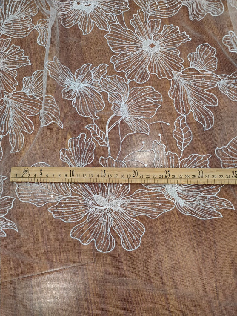 May include: White floral lace fabric with a beaded design. The fabric is laid out on a wooden surface with a ruler measuring 35 inches.