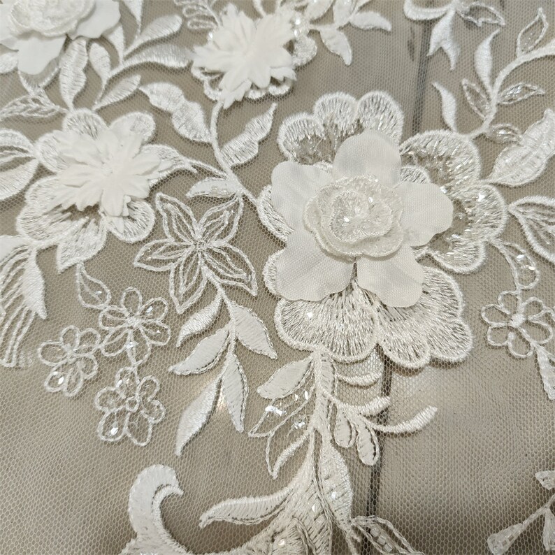 May include: White floral lace fabric with 3D flowers and sequins. The lace is on a sheer mesh background.