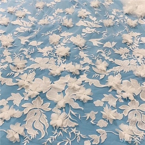 May include: White floral lace fabric with 3D flowers on a light blue background. The lace is delicate and intricate, with a variety of flower shapes and sizes. The fabric is perfect for wedding dresses, bridal accessories, and other special occasion garments.