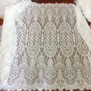 May include: White lace fabric with a delicate, intricate pattern. The lace features a repeating design of arches, circles, and geometric shapes. The fabric has a soft, flowing texture.