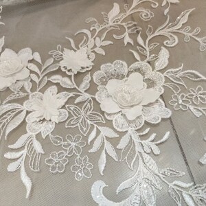 May include: White floral lace fabric with 3D flowers and sequins. The lace is delicate and intricate, with a floral pattern that is both classic and elegant. The fabric is perfect for a variety of projects, including wedding dresses, evening gowns, and home decor.