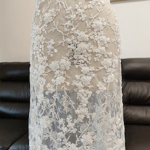 May include: White floral lace fabric with a delicate, embroidered design. The lace features detailed flower and vine patterns, creating a textured, see-through effect. This fabric is suitable for bridal wear, dresses, or other special occasion garments.