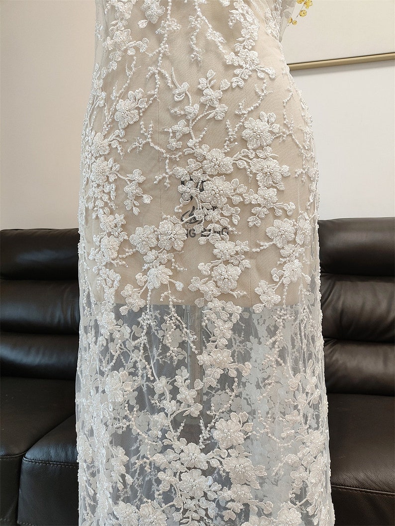 May include: White floral lace fabric with a delicate, embroidered design. The lace features detailed flower and vine patterns, creating a textured, see-through effect. This fabric is suitable for bridal wear, dresses, or other special occasion garments.