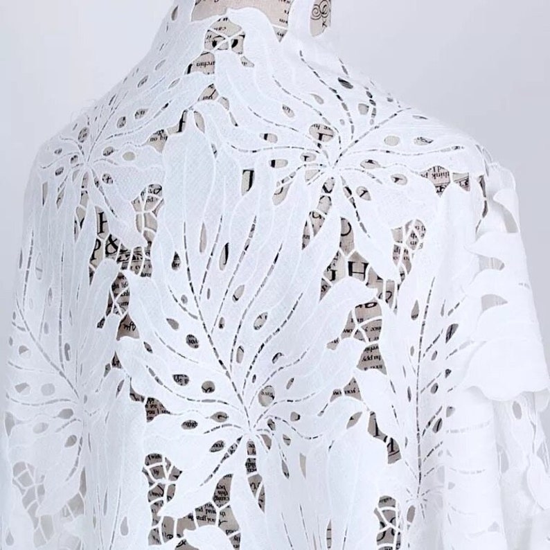 May include: White cotton fabric with a delicate, intricate laser-cut design of large leaves. The fabric is sheer and lightweight, perfect for creating garments or home decor.