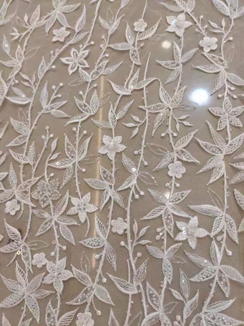 May include: White floral lace fabric with a delicate pattern of flowers and leaves. The fabric has a sheer, lightweight texture and is perfect for wedding dresses, bridal accessories, and other special occasion garments.