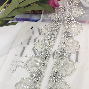 May include: A white tulle trim featuring intricate silver beadwork and clear crystal embellishments arranged in a repeating floral pattern. The decorative border is crafted with metallic seed beads, sequins, and faceted stones on a sheer mesh fabric.