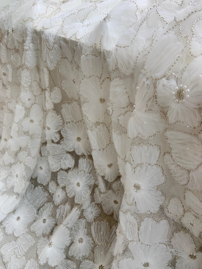May include: White fabric with a delicate floral pattern. The fabric is embellished with small pearls and has a 3D effect.