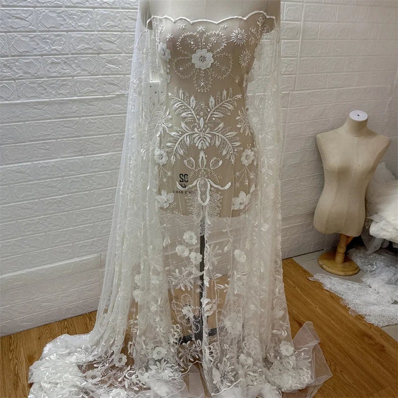 May include: White beaded lace fabric with floral designs and sequins. The fabric is draped over a white mannequin. The fabric is sheer and delicate.