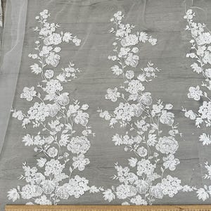 May include: White floral lace fabric with a repeating pattern of flowers and leaves. The fabric is shown against a gray background with a ruler at the bottom of the image.