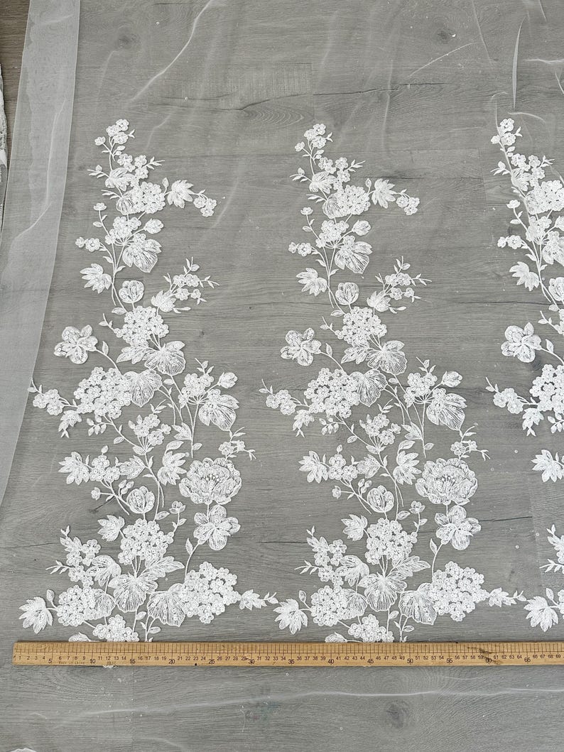 May include: White floral lace fabric with a repeating pattern of flowers and leaves. The fabric is shown against a gray background with a ruler at the bottom of the image.