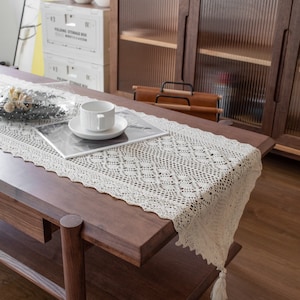 Könnte beinhalten: Cremefarbener Tischläufer aus offener Spitze mit Rautenmuster und Quastendetail. Der Läufer liegt auf einem dunkelbraunen Couchtisch aus Holz. Eine weiße Tasse und Untertasse stehen auf einer Zeitschrift. Der Raum hat eine moderne, minimalistische Ästhetik.