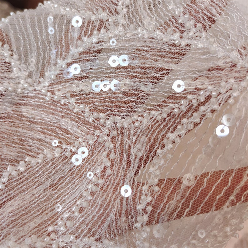 Peut inclure: Tissu blanc transparent avec de petites paillettes et perles blanches. Le tissu a une texture d&eacute;licate et dentel&eacute;e.