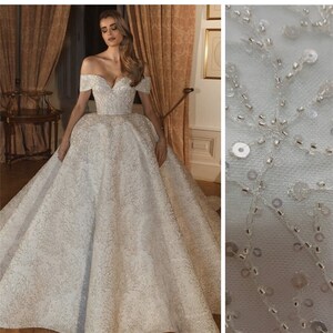 May include: A white off-the-shoulder wedding dress with a full skirt and intricate beading. The dress is made of a soft, flowing fabric and features a sweetheart neckline.
