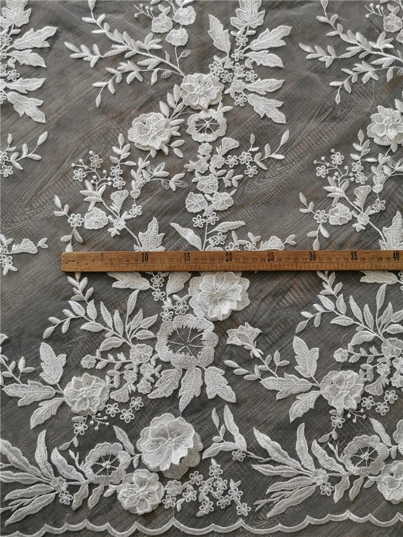 May include: White floral lace fabric with a scalloped edge. The fabric is shown on a wooden surface with a ruler measuring 40 centimeters. The lace has a delicate floral pattern with small and large flowers.