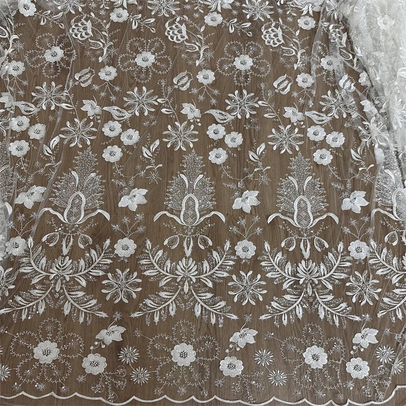 May include: White floral lace fabric with a glittery finish. The lace features a repeating pattern of flowers, leaves, and vines. The fabric is sheer and delicate, and would be perfect for a wedding dress or other special occasion garment.