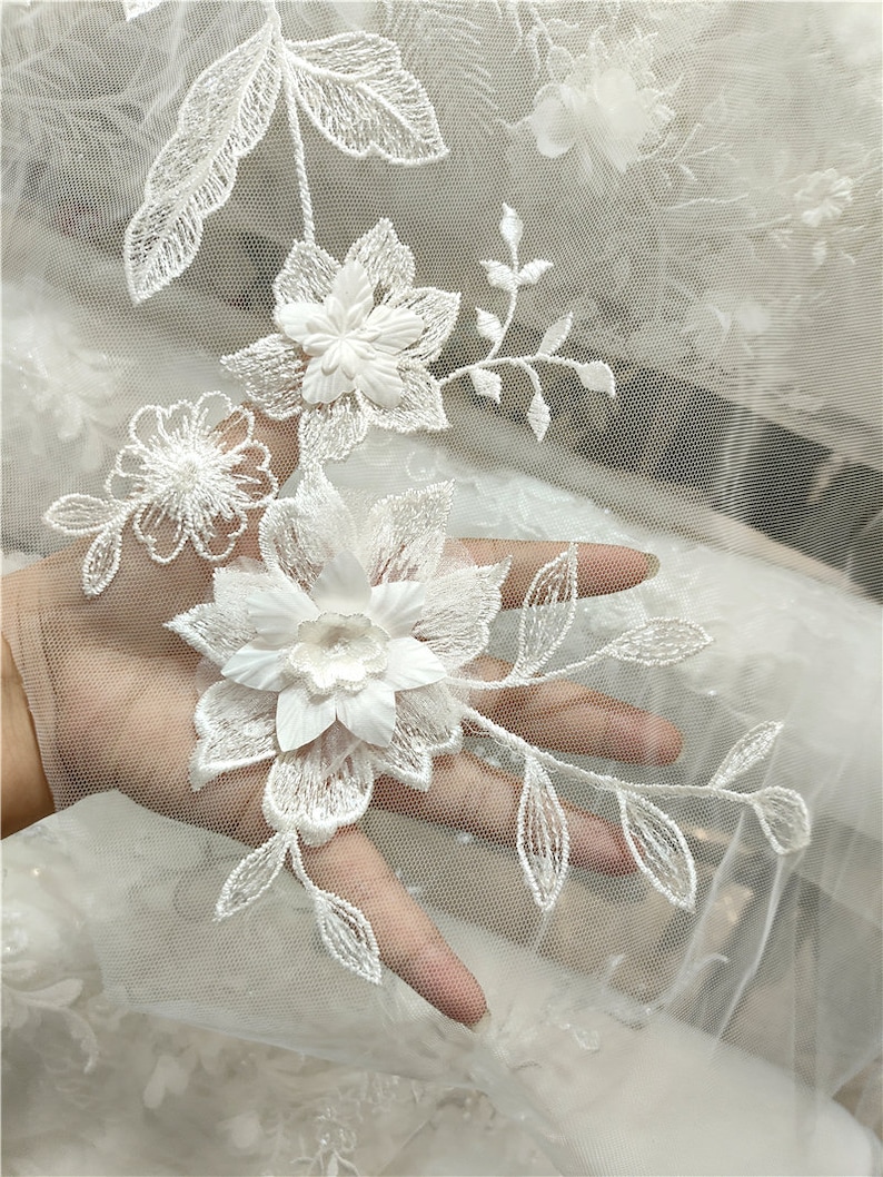May include: White floral lace fabric with delicate embroidery. The lace features intricate floral designs with raised 3D flowers and leaves. The fabric has a soft, sheer texture and is perfect for adding a touch of elegance to any project.