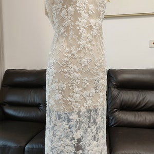 May include: White floral lace fabric draped over a mannequin. The lace features detailed, raised floral embroidery with a delicate, sheer background. The design is densely patterned with flowers and vines, creating a textured, elegant look.