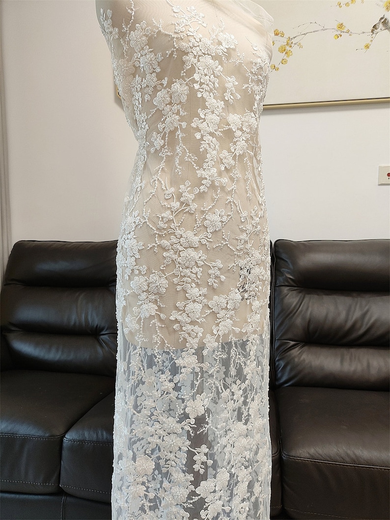 May include: White floral lace fabric draped over a mannequin. The lace features detailed, raised floral embroidery with a delicate, sheer background. The design is densely patterned with flowers and vines, creating a textured, elegant look.