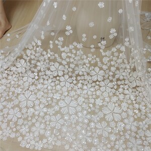 May include: White tulle fabric with white embroidered flowers and sequins. The fabric has a sheer, delicate texture and is perfect for wedding dresses, bridal veils, and other special occasion garments.
