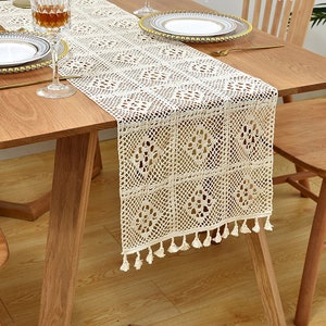 Puede incluir: Un camino de mesa de crochet con un patrón cuadrado y flecos. El camino de mesa es beige y tiene un diseño delicado e intrincado.