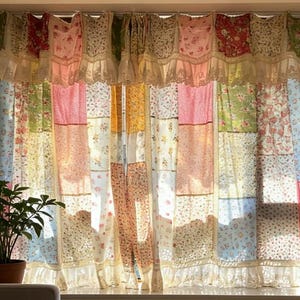 Könnte beinhalten: Ein Patchwork-Vorhangset mit einer Vielzahl von floralen und einfarbigen Stoffquadraten in den Farben Rosa, Blau, Gelb, Grün und Rot. Die Vorhänge bestehen aus einem gerüschten Volant mit Spitzenbesatz und passenden Bahnen vor einem Fenster.