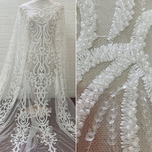 May include: White lace fabric featuring an elaborate floral and scrollwork pattern. The delicate material is adorned with tiny, clear beads, providing texture and shimmer. The lace is draped, highlighting its flowing nature.