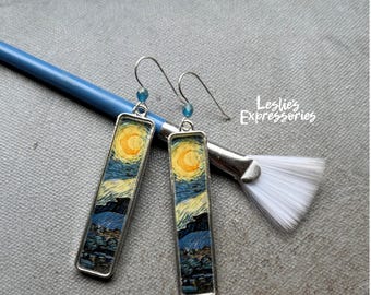 May include: A pair of rectangular drop earrings featuring a printed reproduction of a swirling night sky with a yellow moon, set in silver-toned metal frames with hook wires. The text Leslie's Expressories is visible in white script.