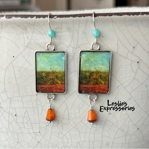 May include: A pair of rectangular earrings with a landscape painting design. The earrings feature a silver frame, a turquoise bead, and an orange teardrop-shaped bead. The landscape painting depicts a field under a blue and yellow sky. The text "Leslie's Expressories" is visible.